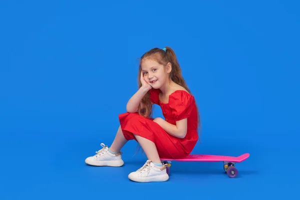 穿着鲜艳衣服的可爱的小女孩坐在滑板上 看着蓝色背景的相机 — 图库照片