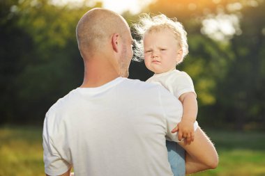 Genç bir baba kucağında sevimli bir bebek tutuyor, arkadan bakıyor. Çocuk üzgün ve gözlerini ovuşturuyor.