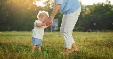 İlk bebek adımları. Genç bir anne parkta çimlerin üzerinde yürümesine yardım eder. Ebeveyn bakımı ve desteği