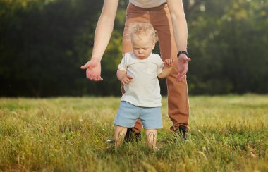 İlk bebek adımları. Genç bir baba, parkta çimlerin üzerinde yürüyen bir çocuğa yardım ediyor. Ebeveyn bakımı ve desteği
