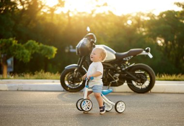Sevimli bebek dengeli bisiklete binmeyi öğreniyor. Küçük bir çocuk arka planda büyük bir motosikletle güneşli bir parkta bisiklet sürmeye çalışıyor. Çocuk motosikletçi olmayı hayal ediyor.
