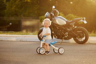 Sevimli bebek dengeli bisiklete binmeyi öğreniyor. Küçük bir çocuk arka planda büyük bir motosikletle güneşli bir parkta bisiklet sürmeye çalışıyor. Çocuk motosikletçi olmayı hayal ediyor.