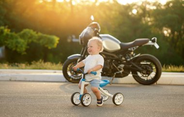 Sevimli bebek dengeli bisiklete binmeyi öğreniyor. Küçük bir çocuk arka planda büyük bir motosikletle güneşli bir parkta bisiklet sürmeye çalışıyor. Çocuk motosikletçi olmayı hayal ediyor.
