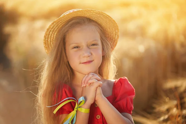 Portrait of a cute baby girl with beautiful long hair in a trendy straw hat. Against the backdrop of a wheat field at sunset. Ukrainian patriot child prays for his country