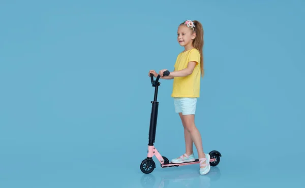 穿着夏装的小女孩骑着滑板车被隔离在蓝色的墙壁背景下 儿童工作室的肖像 童年生活方式概念 — 图库照片