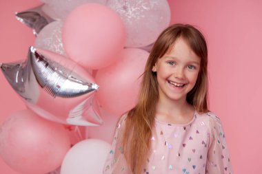 Joyful child girl in elegant tulle dress near the balloons. Birthday present. Funny face celebrates birthday party on pink background	