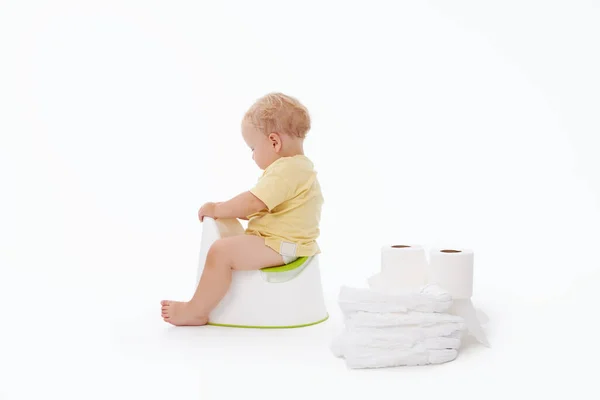 Potty training a baby. Little boy sitting on a potty next to a roll of toilet paper and diapers on a white background