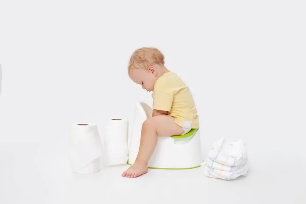 Potty training a baby. Little boy sitting on a potty next to a roll of toilet paper and diapers on a white background