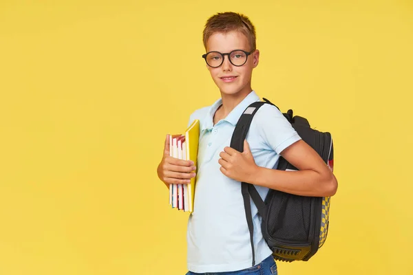 Portrait of a schoolboy in glasses with textbooks and a backpack on a yellow background. Back to school