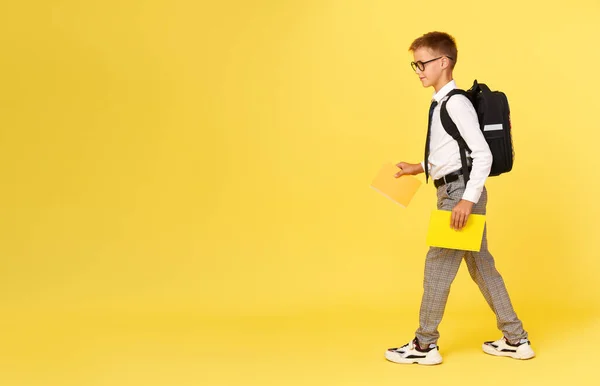 Portrait of a schoolboy in glasses with textbooks and a backpack on a yellow background steps to the left. Back to school