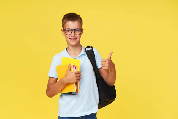Portrait of a schoolboy in glasses with textbooks and a backpack on a yellow background showing ok sign. Back to school