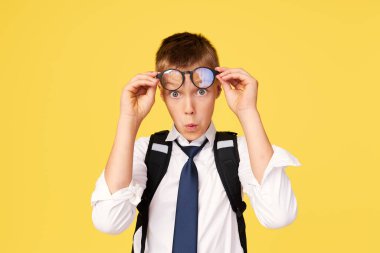 Gözlüklü, ders kitabı ve sarı arka planda sırt çantası olan şaşırmış bir okul çocuğunun portresi. Okula geri dön.