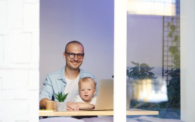 Genç baba kucağında küçük oğluyla uzaktan kumandayla çalışıyor.