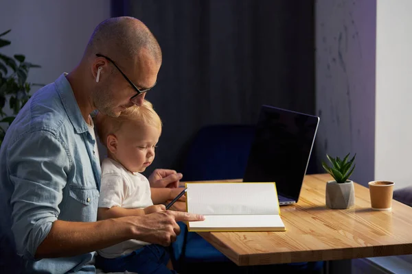 Young father working remotely on laptop with little baby son in his arms