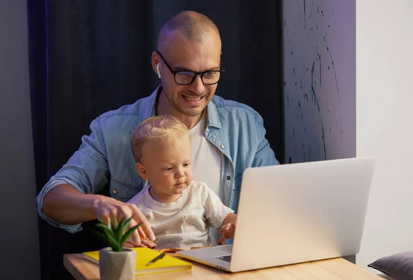 Young father working remotely on laptop with little baby son in his arms