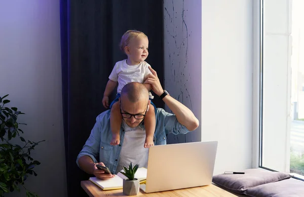 Young father working remotely on laptop with little baby son in his arms