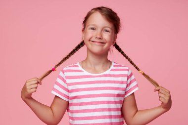 Portrait of surprised cute  toddler girl child standing isolated over pink background. holding hands on two pigtails. Looking at camera. 