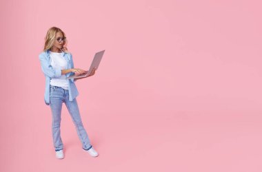 Portrait of a young girl in casual denim blue clothes with a laptop in her hands on a pink background. The concept of learning and job search online