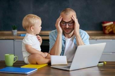 Young father freelancer working on laptop remotely from home office with baby in his arms. Concept of resilience while working from home with kids during quarantine