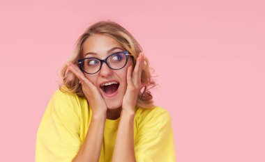Portrait of an excited young woman in eyeglasses, yellow t-shirt on a pink background. Surprised girl looks to the right with her eyes wide open. Wow face