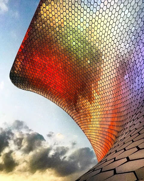 colored light reflected on the latticework of the museum, with clouds and sky in the background, reds, greens, oranges and purples projected on the museum.