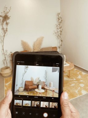 young woman taking a picture of a room, two armchairs, coffee table, space with carpet and white painted walls.