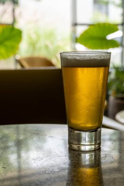 glass of beer on a terrazzo table, two yellow chairs in the background, retro table, window with natural light and potted plants, white painted brick, white painted brick