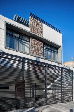 facade of a modern house with stone, staple that wraps the windows painted in gray, metal entrance, ironwork, as well as the terrace in the same material, two large windows on the facade.