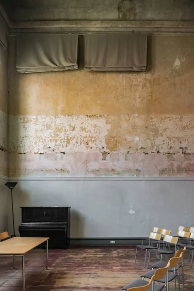 grand piano, or normal piano, in a vintage room, unpainted walls, with dirt and humidity, wooden chairs and desk, classroom or music room, with lamp in a corner, without people.