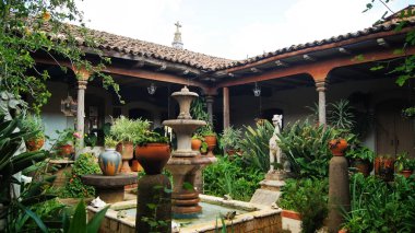 stone fountain, in old mexican house, latin america, with decorated tiles, roof tiles, surrounding vegetation and pots, aged materials.
