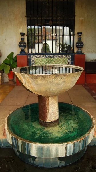 stone fountain, in old mexican house, latin america, with decorated tiles, roof tiles, surrounding vegetation and pots, aged materials.