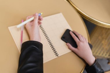 Yuvarlak masa üzerinde boş defterin yanında elinde kalem ve akıllı telefon tutan kadının kırpılmış görüntüsü 