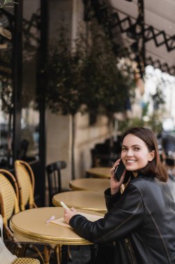 Deri ceketli neşeli kadın akıllı telefonla konuşuyor ve Fransa 'da açık bir kafede oturuyor.