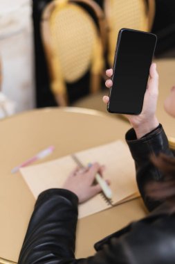 Yuvarlak masadaki bulanık defterin yanında elinde boş ekranla akıllı telefon tutan bir kadın görüntüsü.