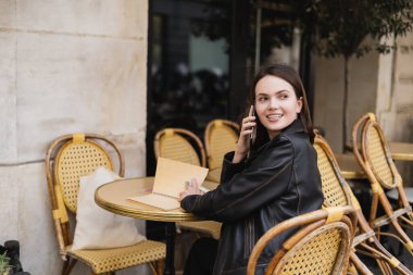 Deri ceketli gülümseyen kadın akıllı telefondan konuşuyor ve açık kafe terasında oturuyor.