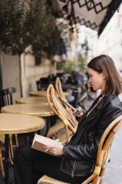Deri ceketli, elinde cep telefonu olan ve açık kafe terasında oturan kadının yan görüntüsü.