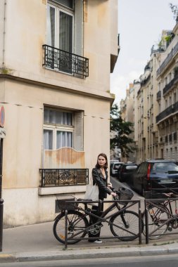 Siyah deri ceketli genç şık bir kadın Paris 'te sokakta bisikletin yanında duruyor. 