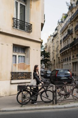 Siyah deri ceketli, şık bir kadın Paris 'te bisikletin yanında dikiliyor. 