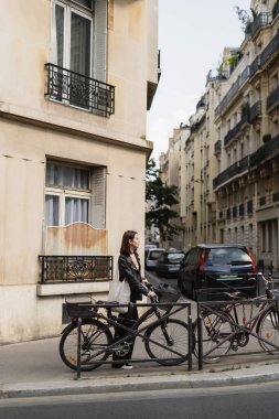 side view of young stylish woman in black leather jacket standing near bicycle on street in paris  clipart