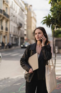 Siyah deri ceketli genç serbest yazar dizüstü bilgisayar tutuyor ve Paris 'te bir sokakta akıllı telefondan konuşuyor.
