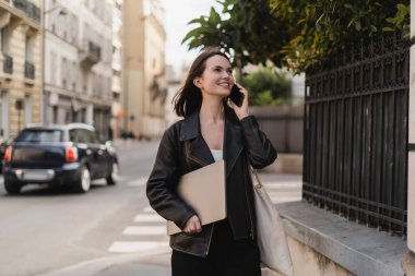 Siyah deri ceketli neşeli serbest yazar dizüstü bilgisayar tutuyor ve Paris 'te bir sokakta akıllı telefondan konuşuyor.