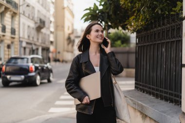 cheerful freelancer in black leather jacket holding laptop and talking on smartphone on street in paris clipart