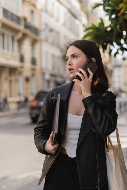 Siyah deri ceketli genç serbest yazar dizüstü bilgisayar tutuyor ve Paris 'te bir sokakta cep telefonuyla konuşuyor.