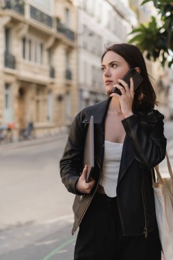 Siyah deri ceketli genç bir kadın dizüstü bilgisayar tutuyor ve Paris 'te bir sokakta akıllı telefondan konuşuyor.