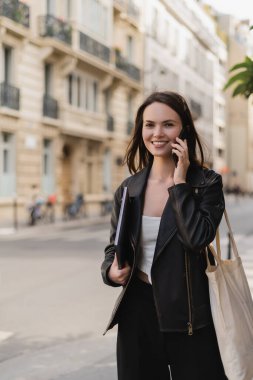 Siyah deri ceketli neşeli bir kadın dizüstü bilgisayar tutuyor ve Paris 'te bir sokakta akıllı telefondan konuşuyor.