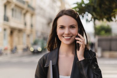Siyah deri ceketli mutlu bir kadının portresi dizüstü bilgisayar tutuyor ve Paris 'te bir sokakta akıllı telefondan konuşuyor.