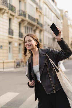 Siyah deri ceketli mutlu kadın dizüstü bilgisayar tutuyor ve Paris 'te sokakta akıllı telefonuyla el sallıyor.