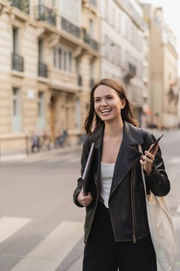 Siyah deri ceketli neşeli bir kadın dizüstü bilgisayar ve akıllı telefonu Paris sokaklarında tutuyor.