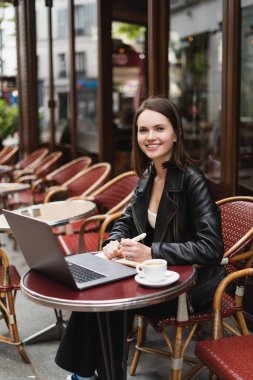 Siyah ceketli mutlu bir serbest yazar laptopun yanındaki kameraya ve açık hava kafedeki masada bir fincan kahveye bakıyor.