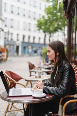 Siyah ceketli, gülümseyen serbest çalışan açık hava kafede kahve masasının yanındaki laptopa bakıyor. 